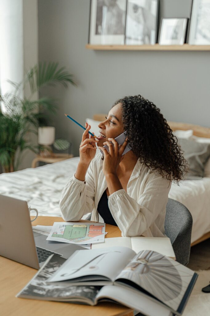 African American woman multitasking at home desk with laptop and phone, embodying modern remote work life.