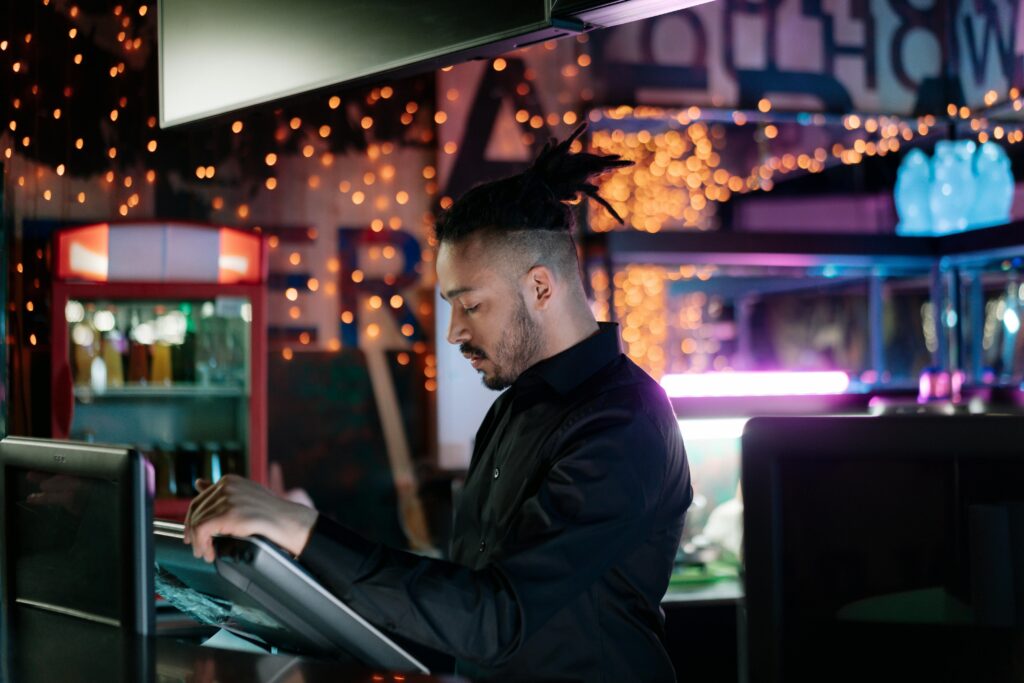 A man with facial hair operates a computer in a trendy bar with festive lights.